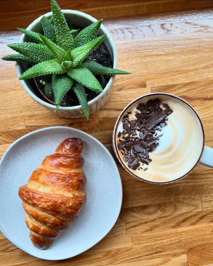 A croissant beside a flat white with chocolate shavings, on the wooden counter