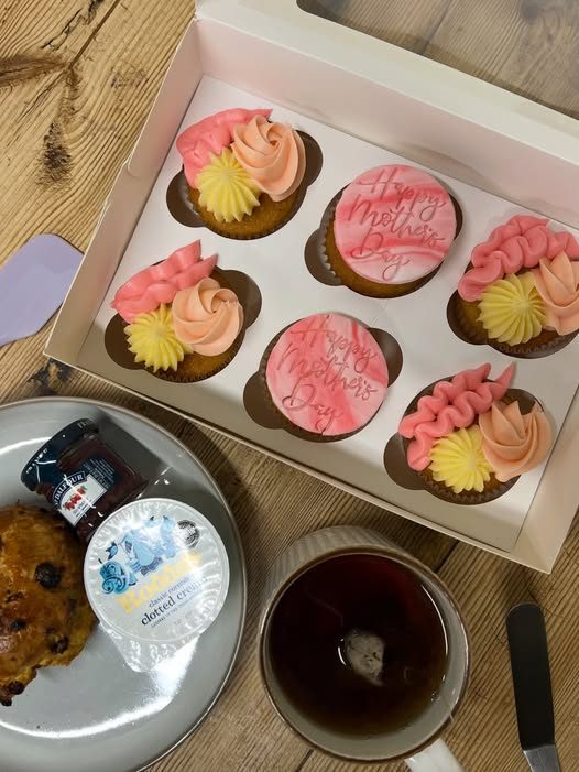 A box of six piped cupcakes beside a cookie plate and coffee