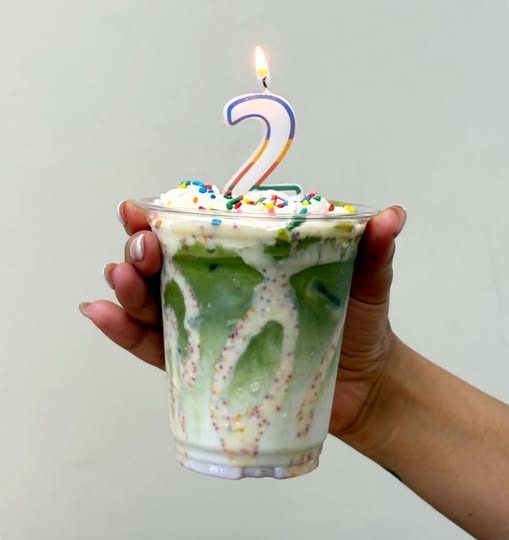 An iced birthday matcha held up with a lit number-two candle on top